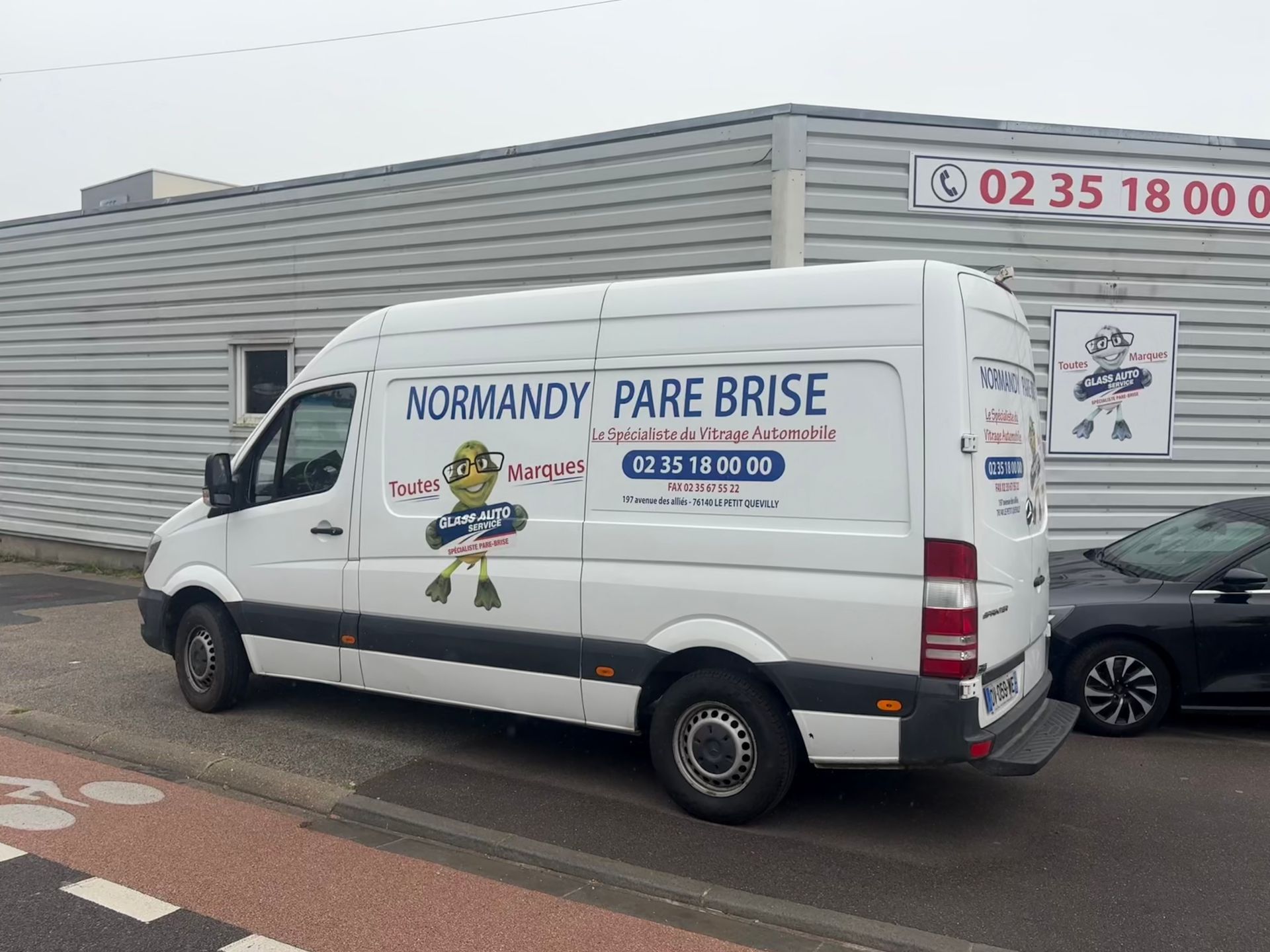 Une camionnette de Normandy Pare-Brise est garée à côté d'un bâtiment avec des enseignes, et une voiture se trouve à proximité.