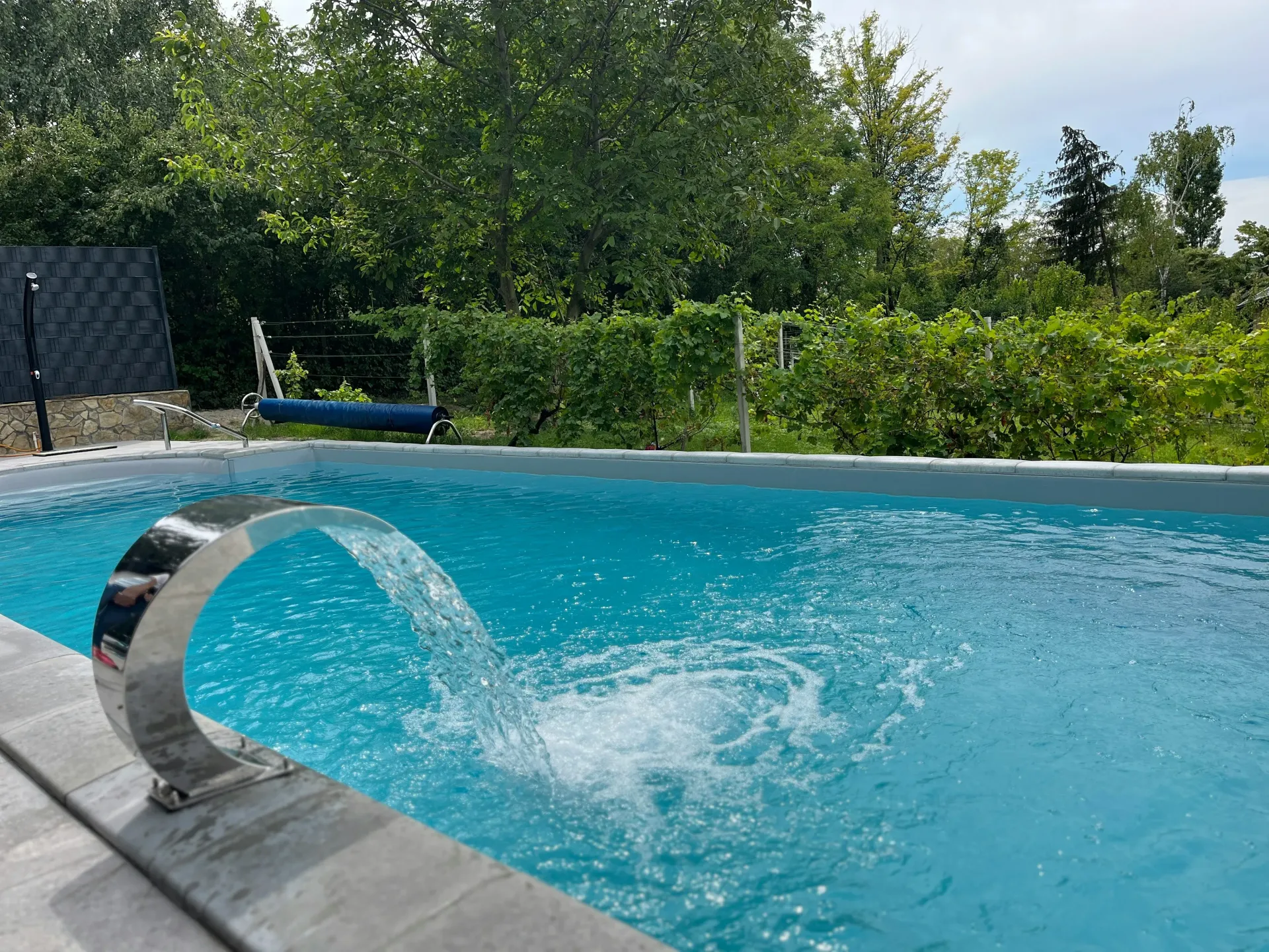 Piscina con cascada, agua azul, cubierta gris y exuberante vegetación de fondo.