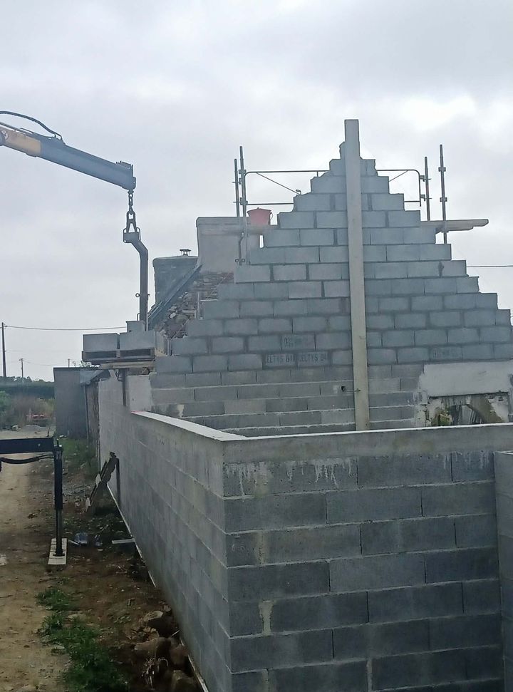 Maison en parpaings partiellement construite, avec toit à gradins et échafaudages à côté.