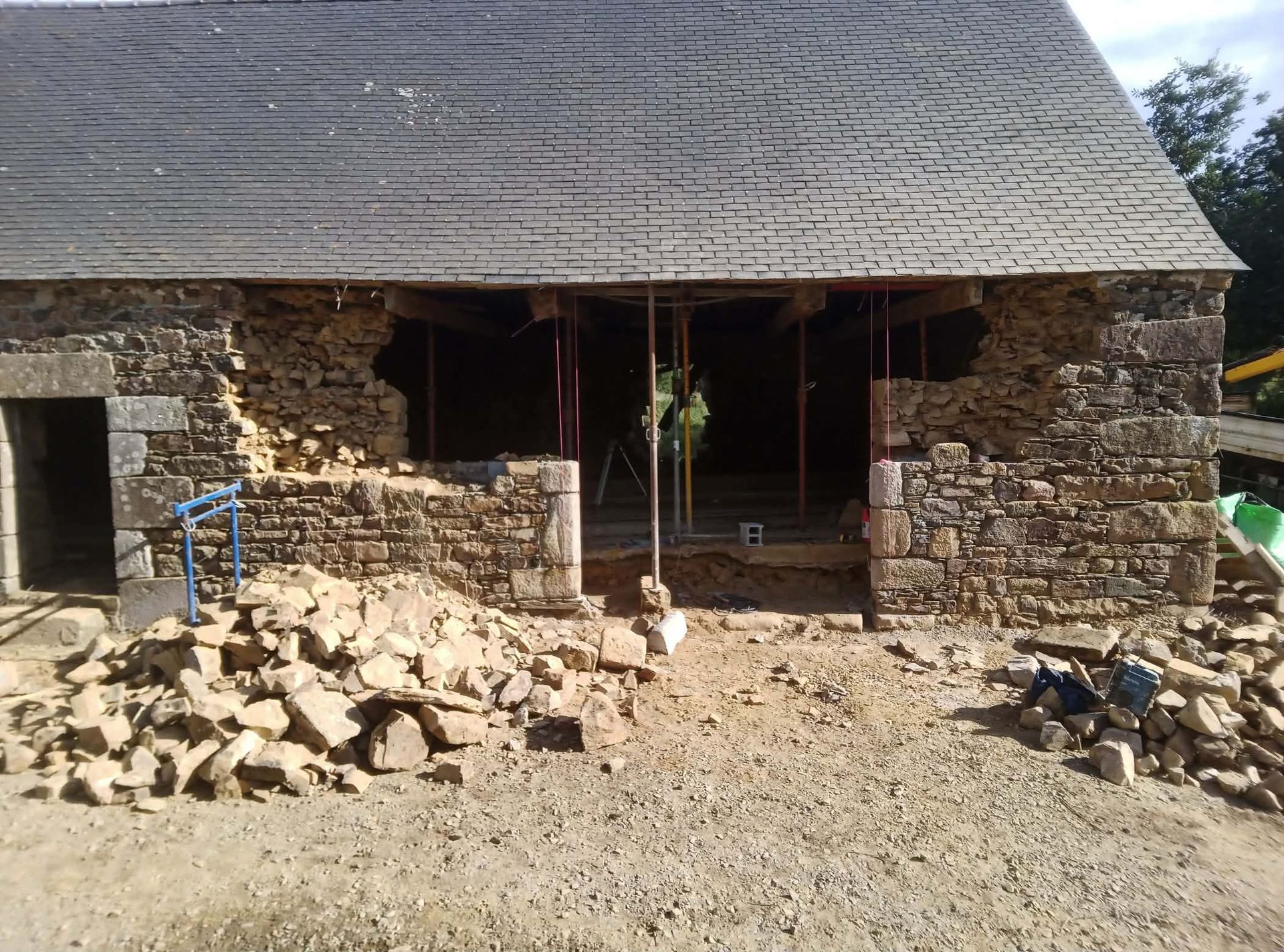 Maison en pierre au toit et au porche inachevé, entourée de gravats et de terre.