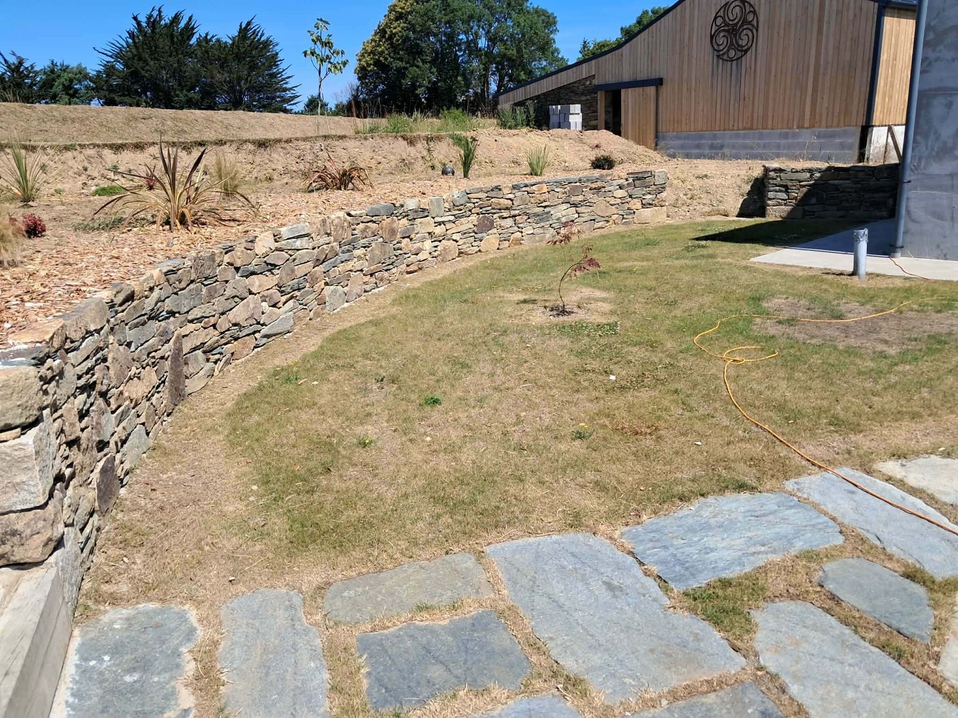 Mur de soutènement en pierre jouxtant une pelouse sèche et une terrasse en dalles.