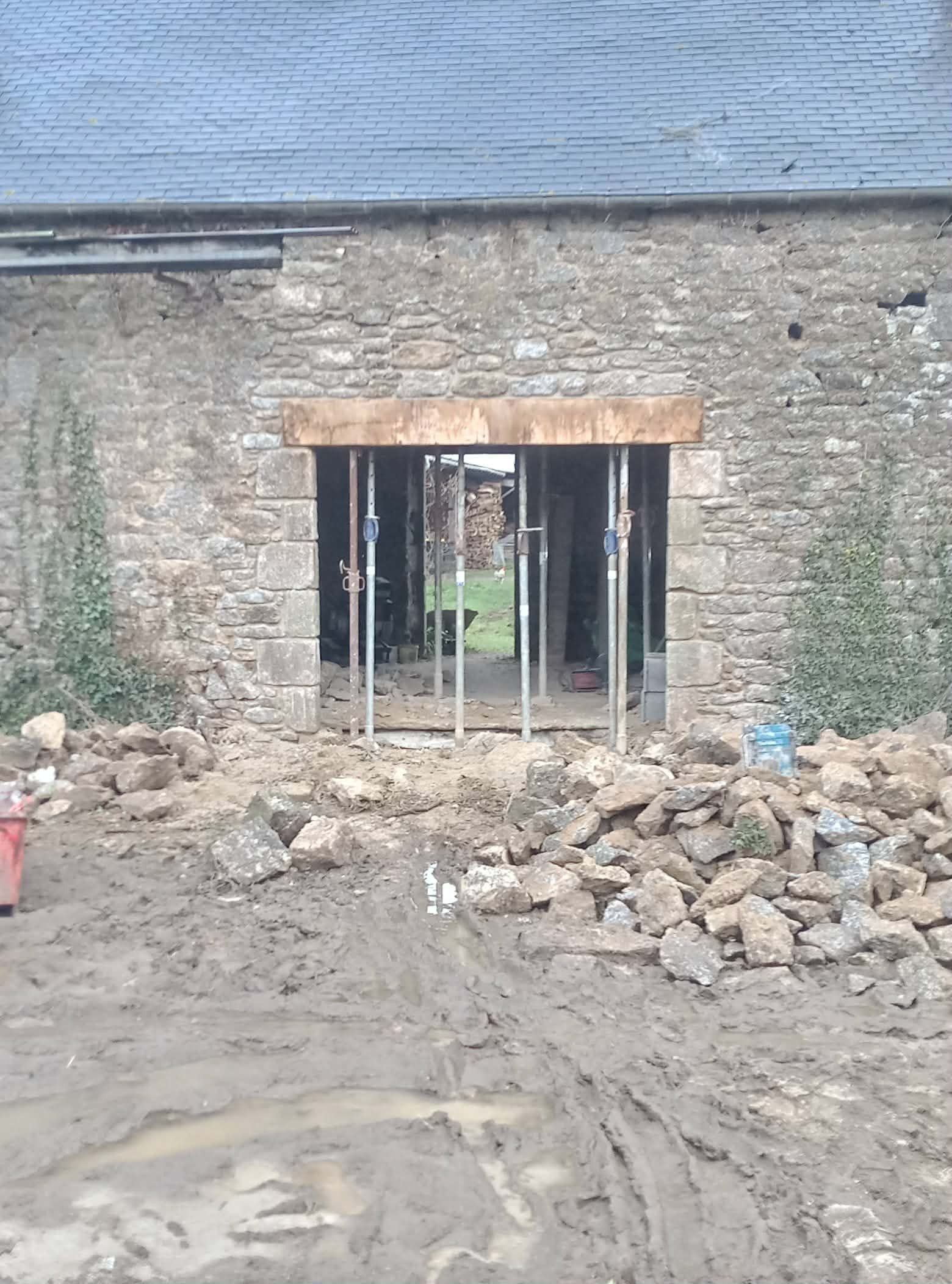 Bâtiment en pierre inachevé avec une ouverture pour une porte, des poteaux de soutien apparents et des gravats devant.