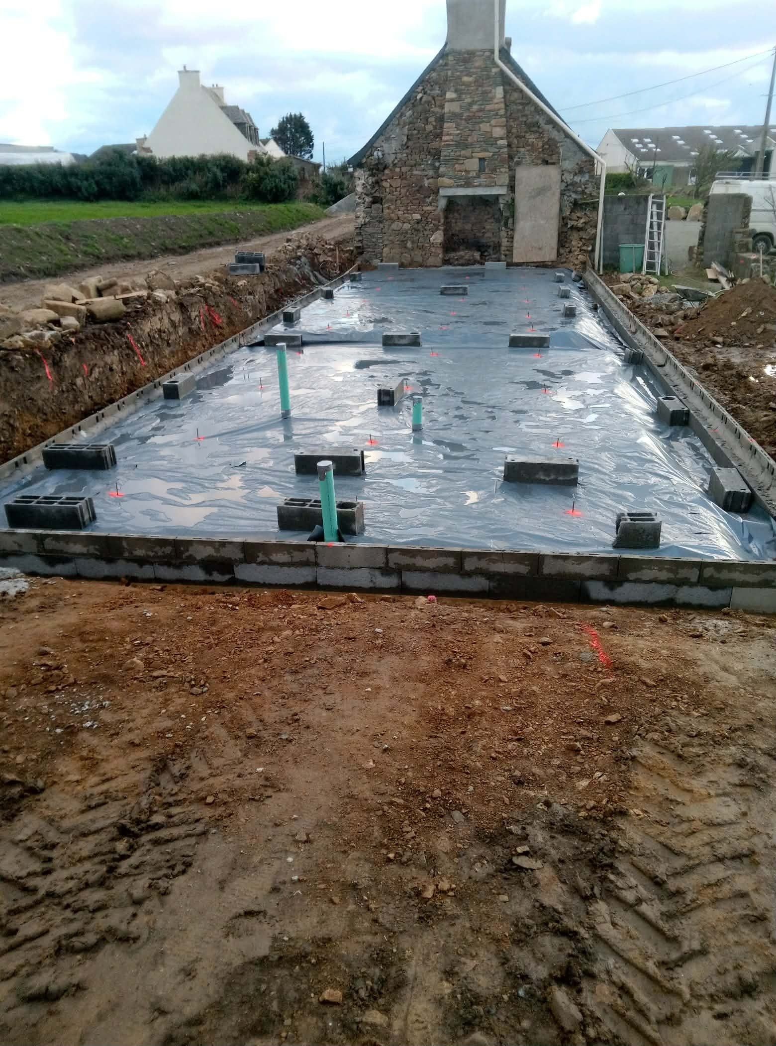 Fondation en dalle de béton partiellement construite sur un chantier rural, avec un bâtiment en pierre en arrière-plan.