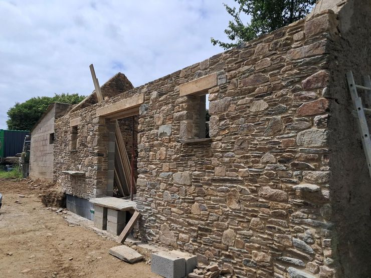 Maison en pierre en construction, avec des murs en maçonnerie brute, une porte en bois et un chantier en terre.