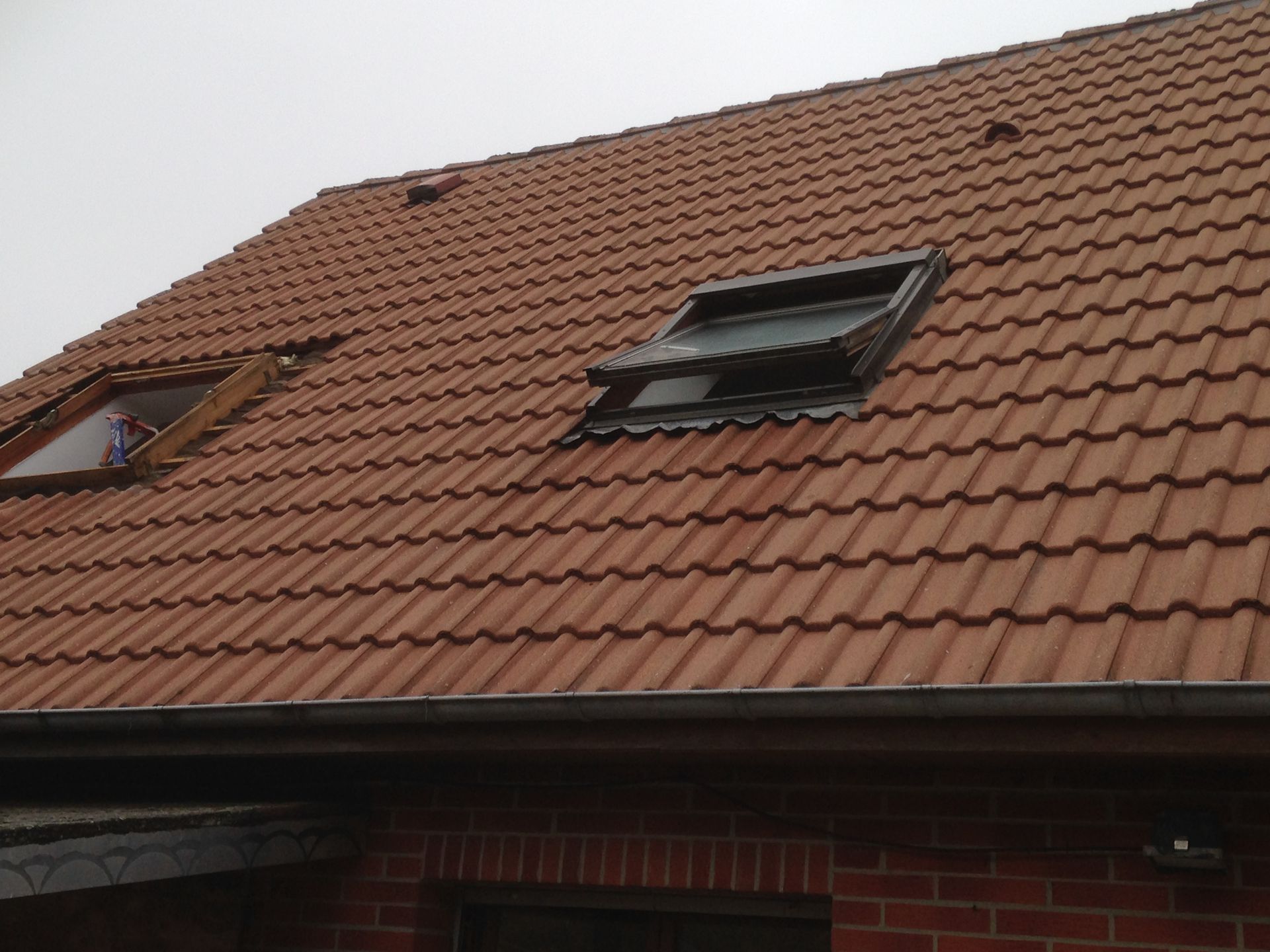 Toit, en tuiles rouges, percé de deux lucarnes. Façade en briques avec encadrements bruns. Ciel nuageux en arrière-plan.