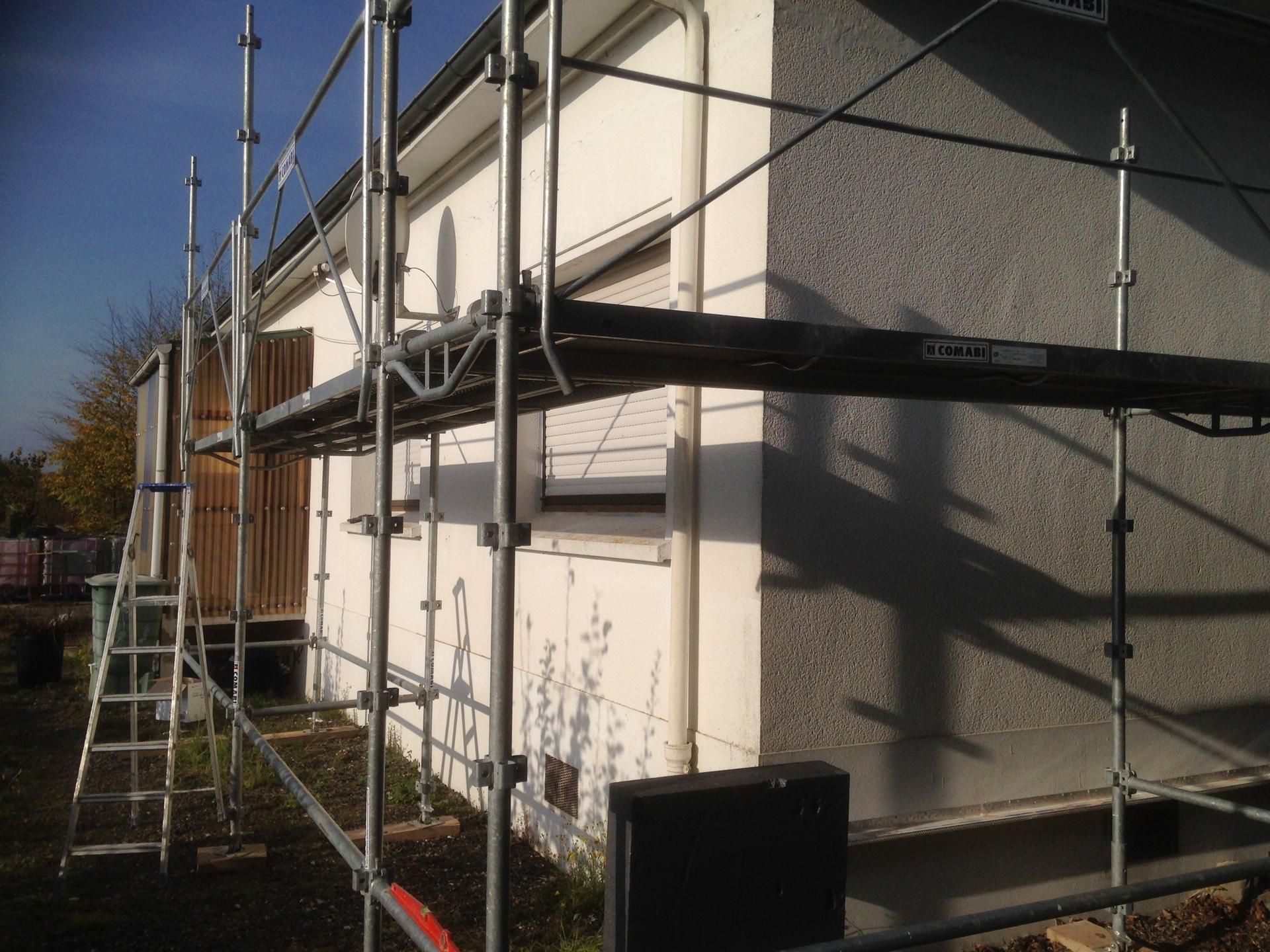 Des échafaudages sont installés le long d'une maison blanche au mur texturé, sous un ciel dégagé.