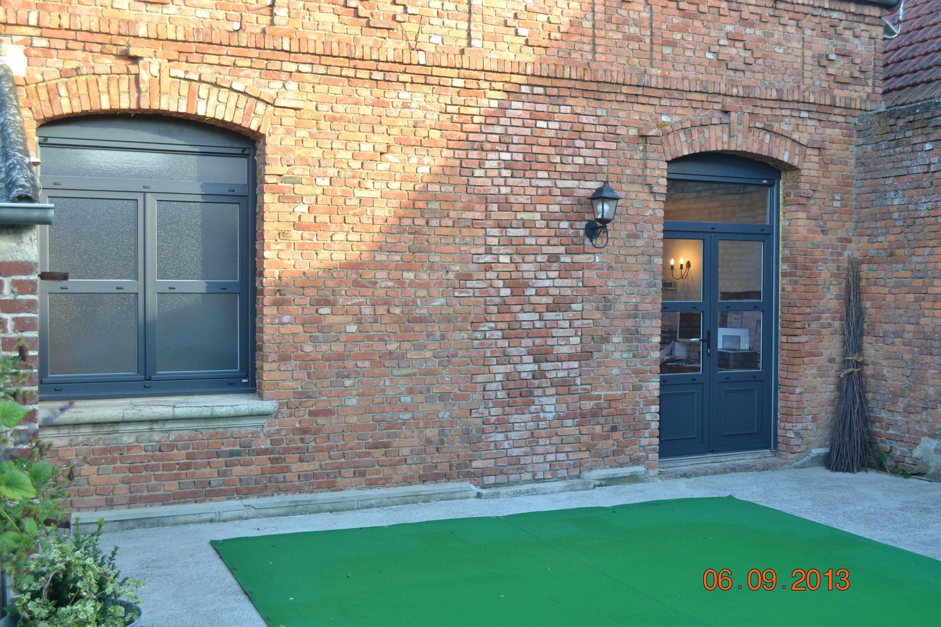 Façade en briques avec une fenêtre et une porte à cadre sombre ; sol recouvert de verdure.