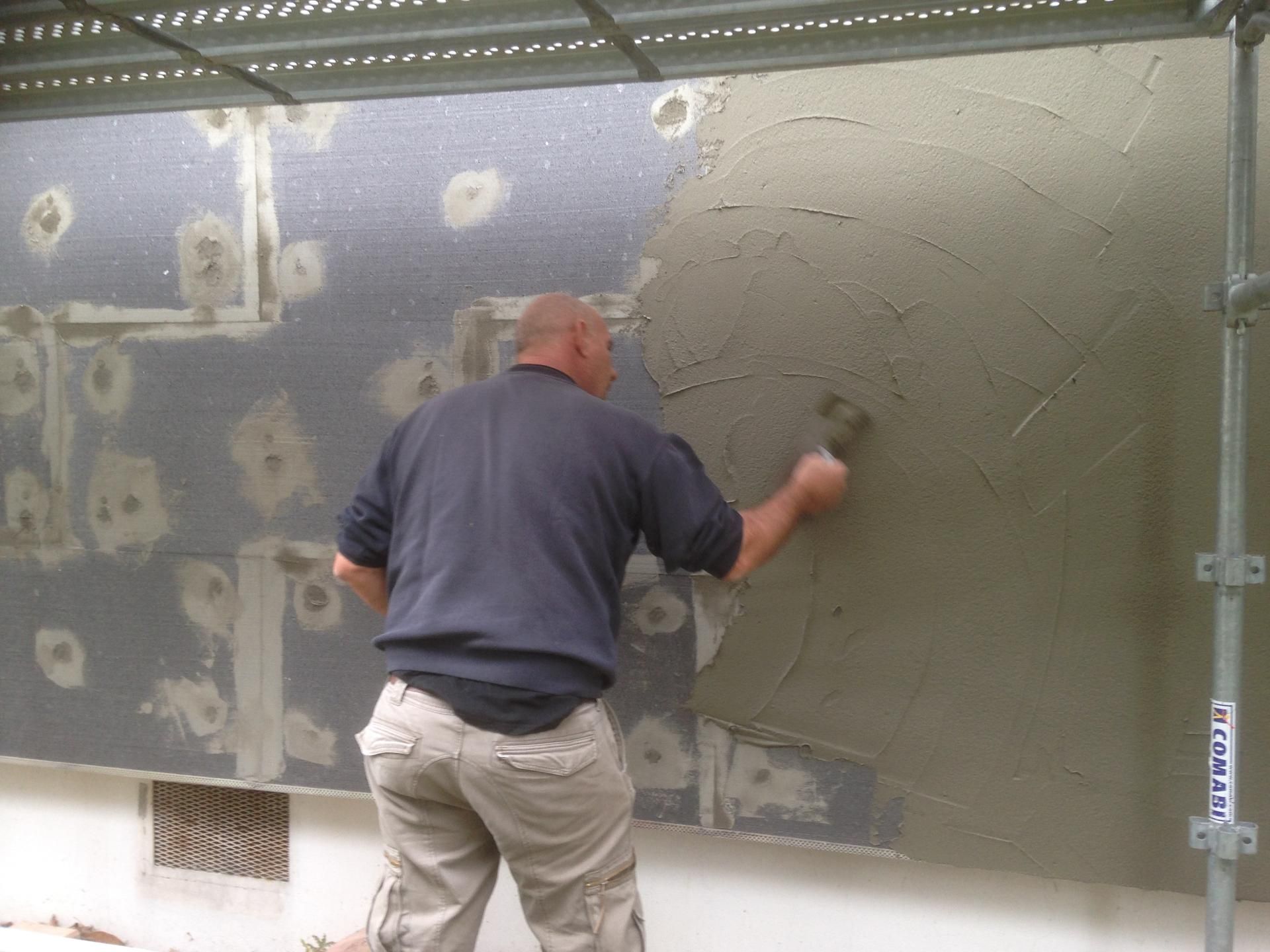 Un homme applique du stuc sur un mur extérieur. L'isolant gris est visible. À l'extérieur, en plein jour.