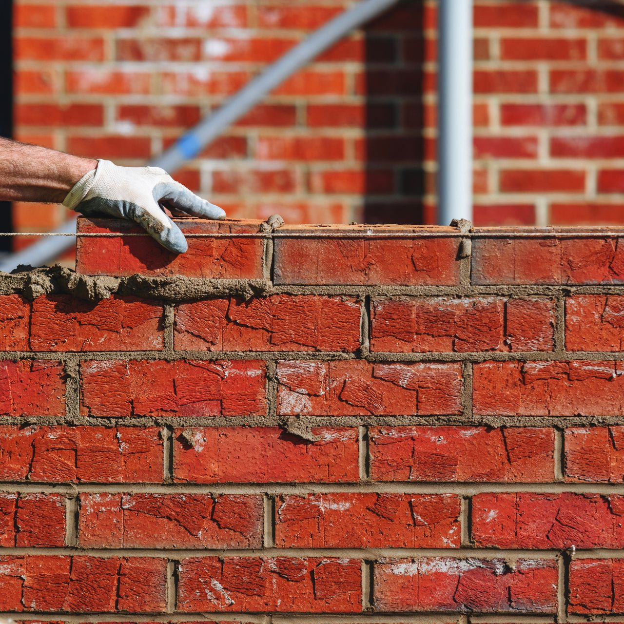 La main d'un maçon gantée désigne la rangée supérieure de briques rouges posées sur un mur, le mortier étant visible.