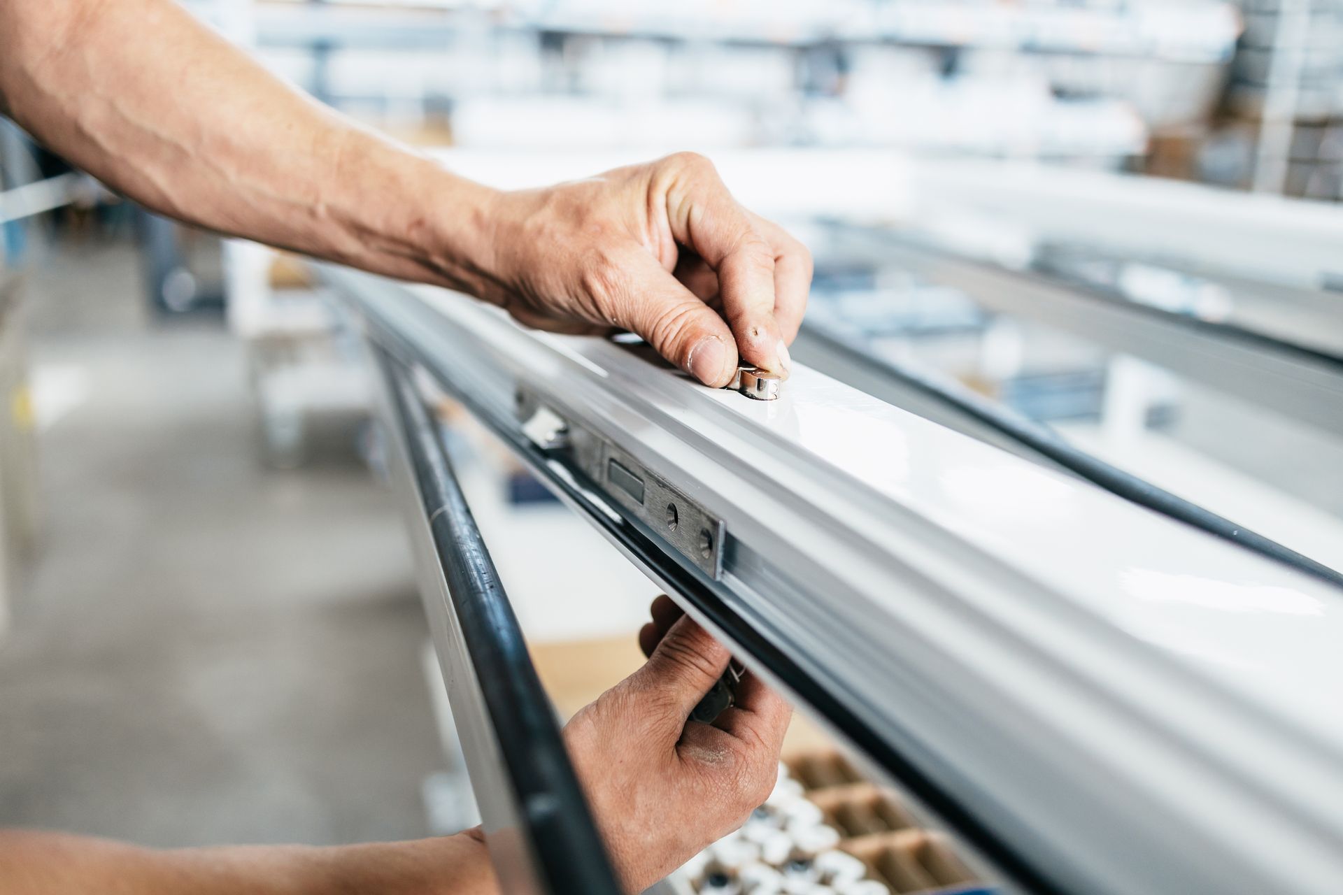 Des mains installent des ferrures sur un cadre de fenêtre métallique en usine.