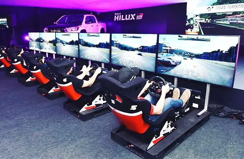 Fila de simuladores de carreras con pantallas grandes en una exhibición de videojuegos, que incluye una Hilux morada.