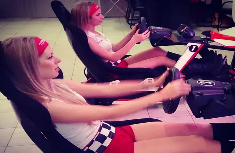Dos mujeres en asientos de carreras usando volantes de videojuegos en un simulador de conducción.
