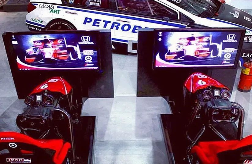 Dos coches de carreras rojos de Fórmula 1 en exhibición con grandes monitores y una pancarta de PETRONAS al fondo.