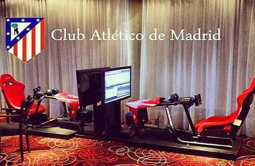 Logotipo del Club Atlético de Madrid en una pantalla junto a simuladores de carreras en una habitación separada por cortinas.
