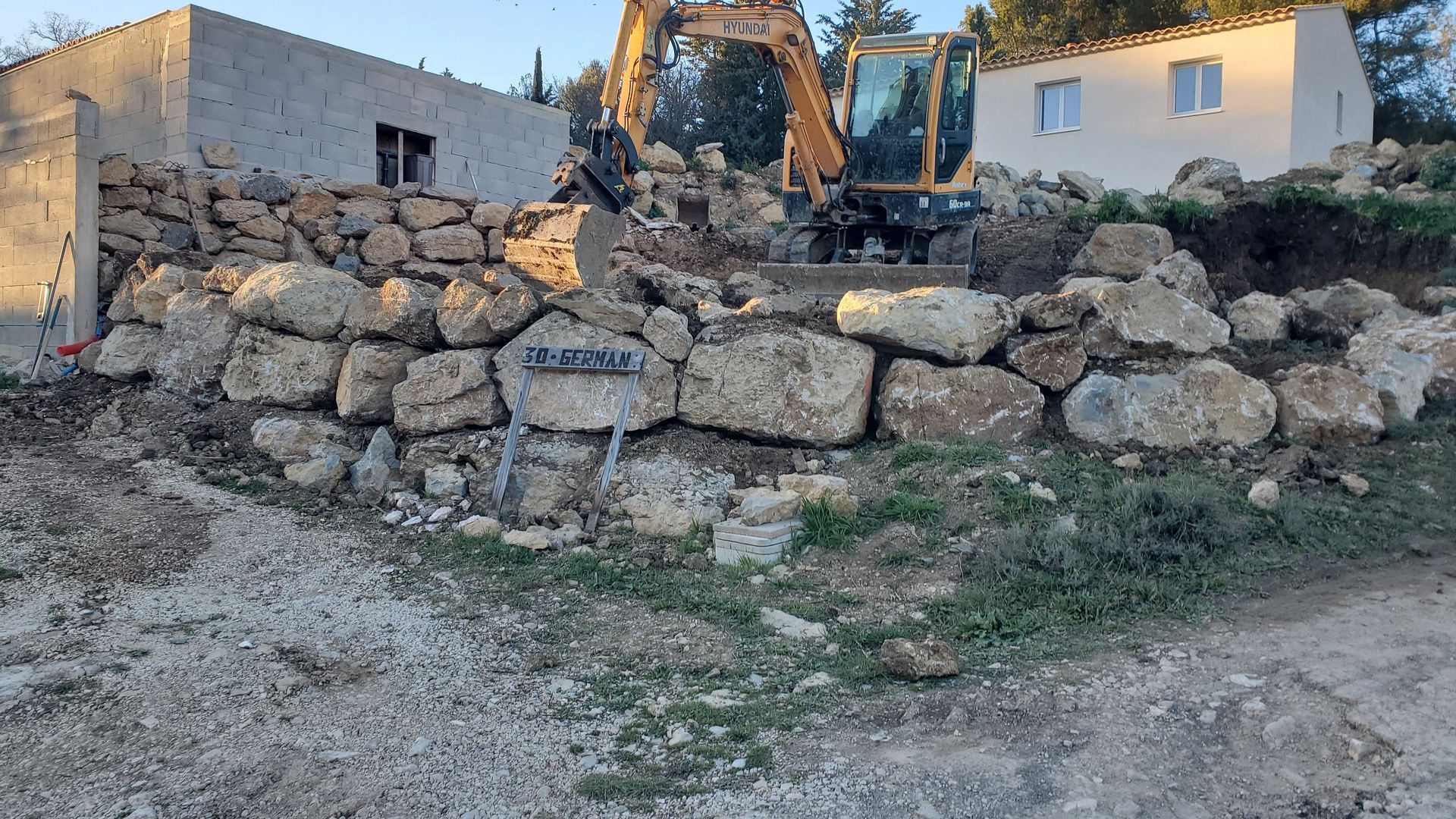 Allée de graviers bordée de verdure et d'un muret, menant vers une montagne au loin.