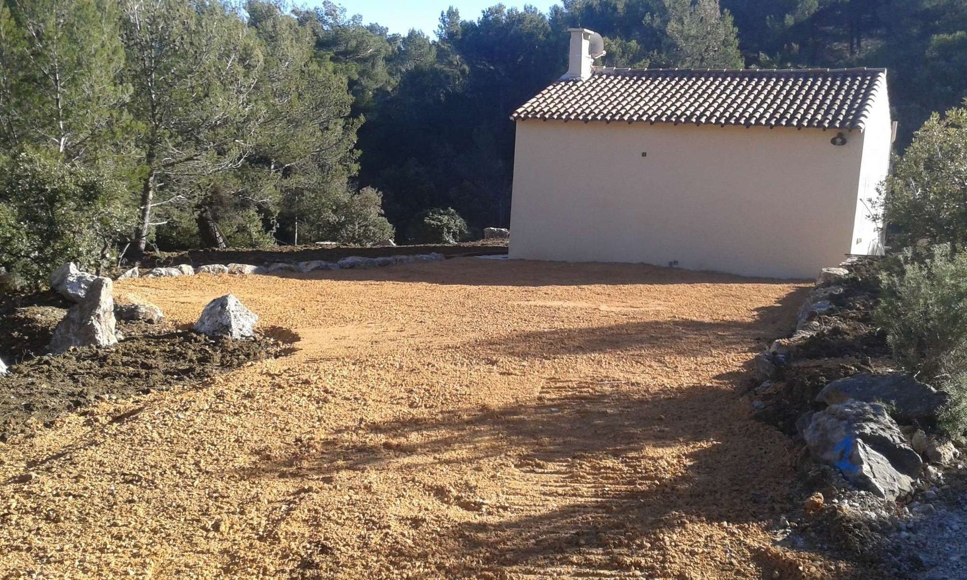 Un petit bâtiment beige au toit de tuiles se dresse au milieu d'une clairière caillouteuse entourée d'arbres.