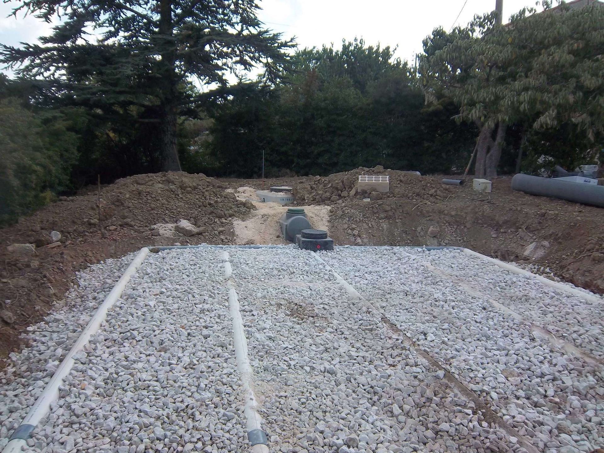 Champ d'épandage en gravier en construction, avec tuyaux perforés et fosse septique, entouré de terre et d'arbres.