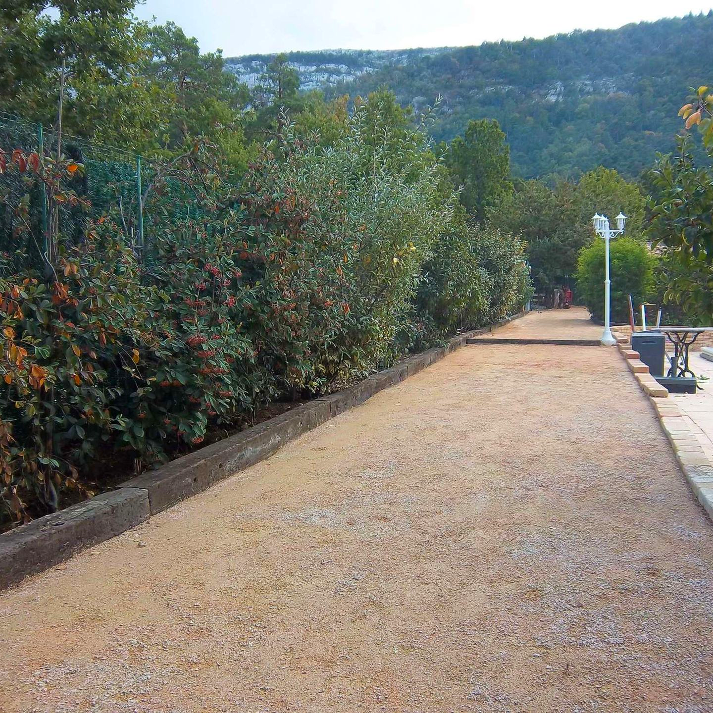 Chemin de gravier bordé de buissons verts et d'un muret, menant à un point de vue panoramique avec des arbres et des collines en arrière-plan.