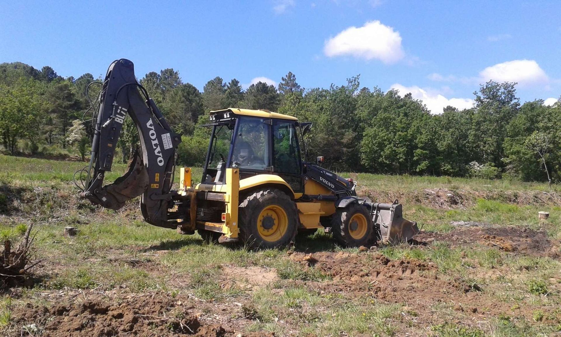 Une pelleteuse Volvo jaune creuse un champ herbeux sous un ciel bleu.