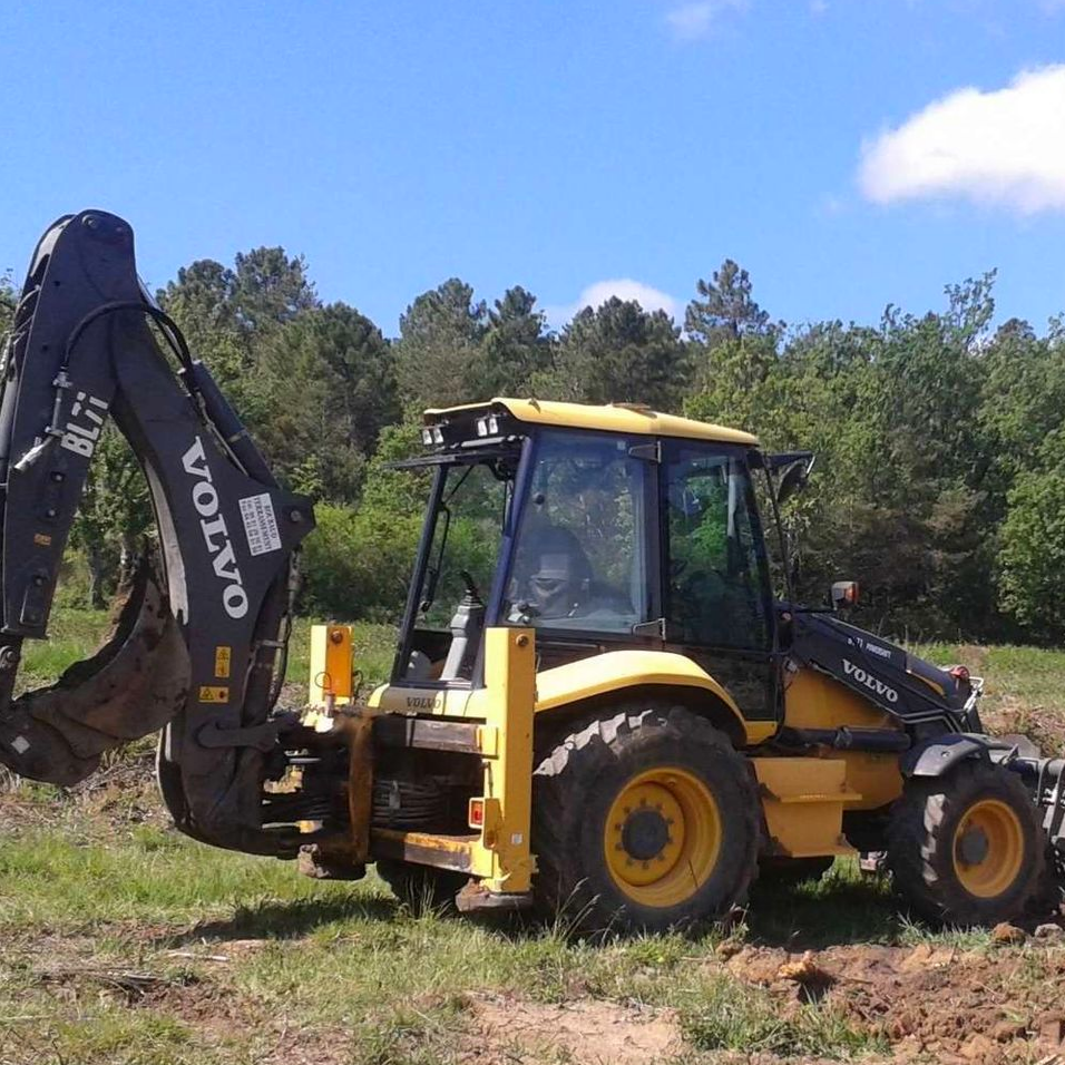 Une pelleteuse Volvo jaune se trouve dans un champ, prête à creuser. Ciel dégagé et arbres en arrière-plan.