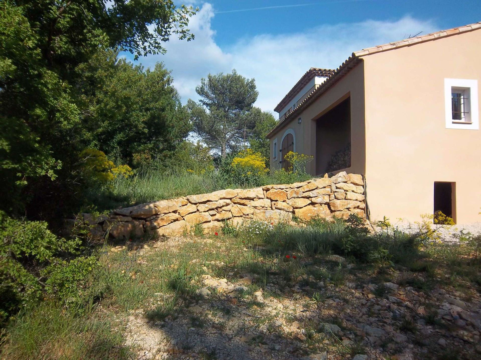 Une maison beige avec un mur de pierre en façade, par une journée ensoleillée. On aperçoit un petit arbre et des plantes en pot.