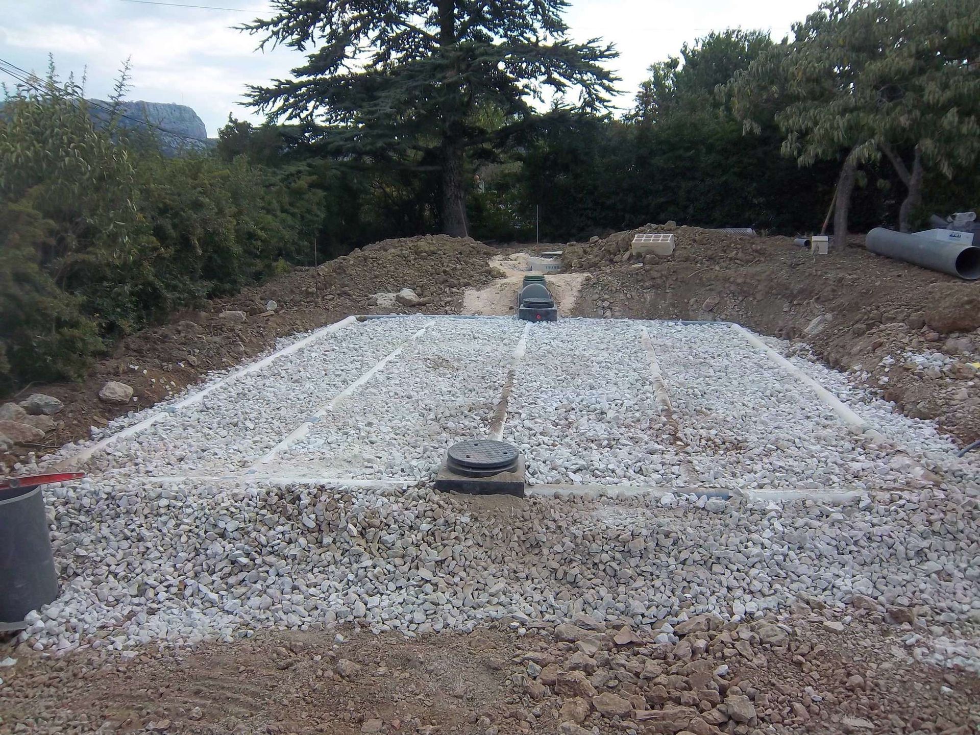Zone de drainage remplie de gravier avec un tuyau central, entourée de terre, arbres en arrière-plan, ciel nuageux.
