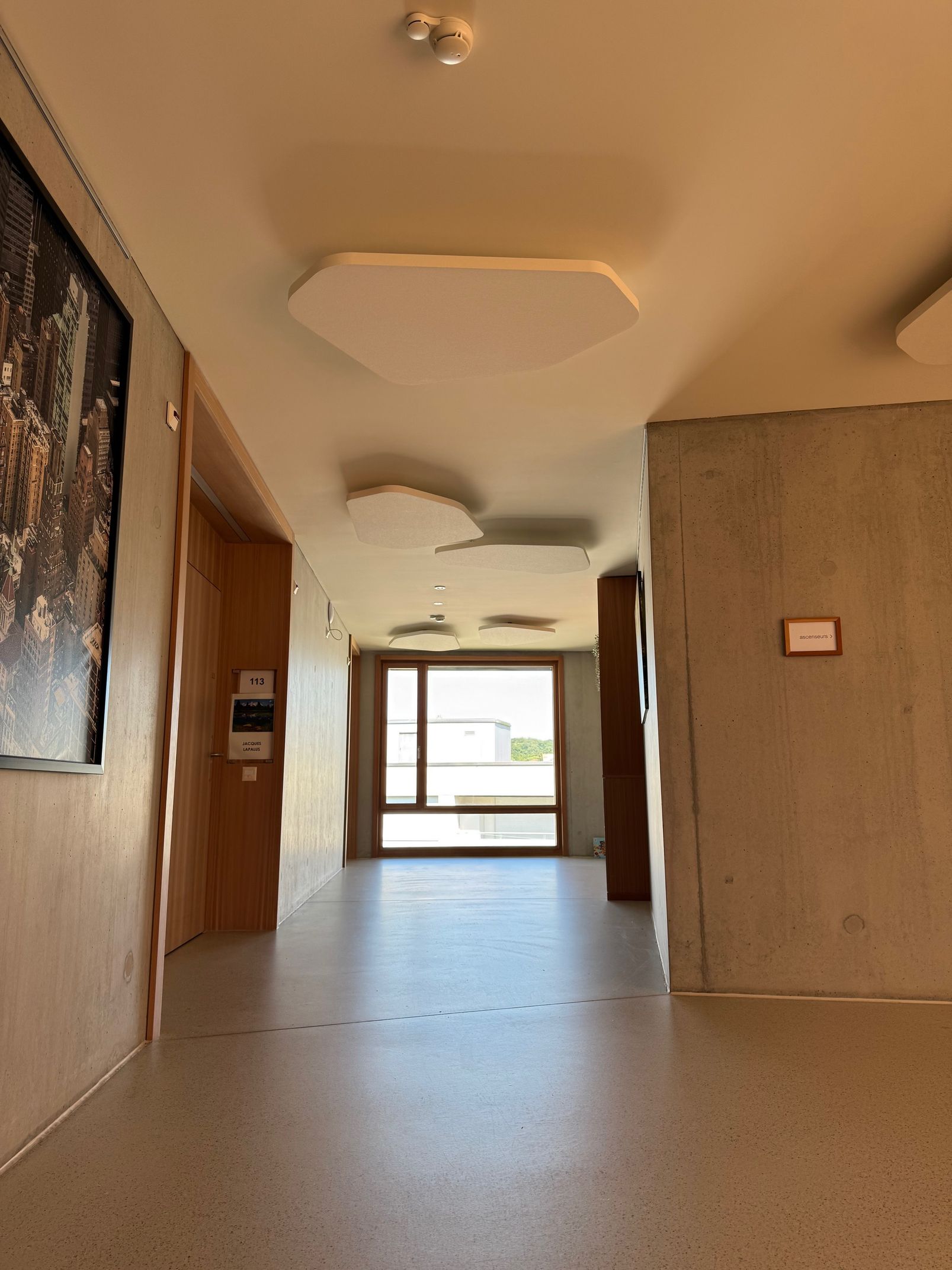 Couloir intérieur vide avec murs beiges, sol carrelé et porte lumineuse au fond.