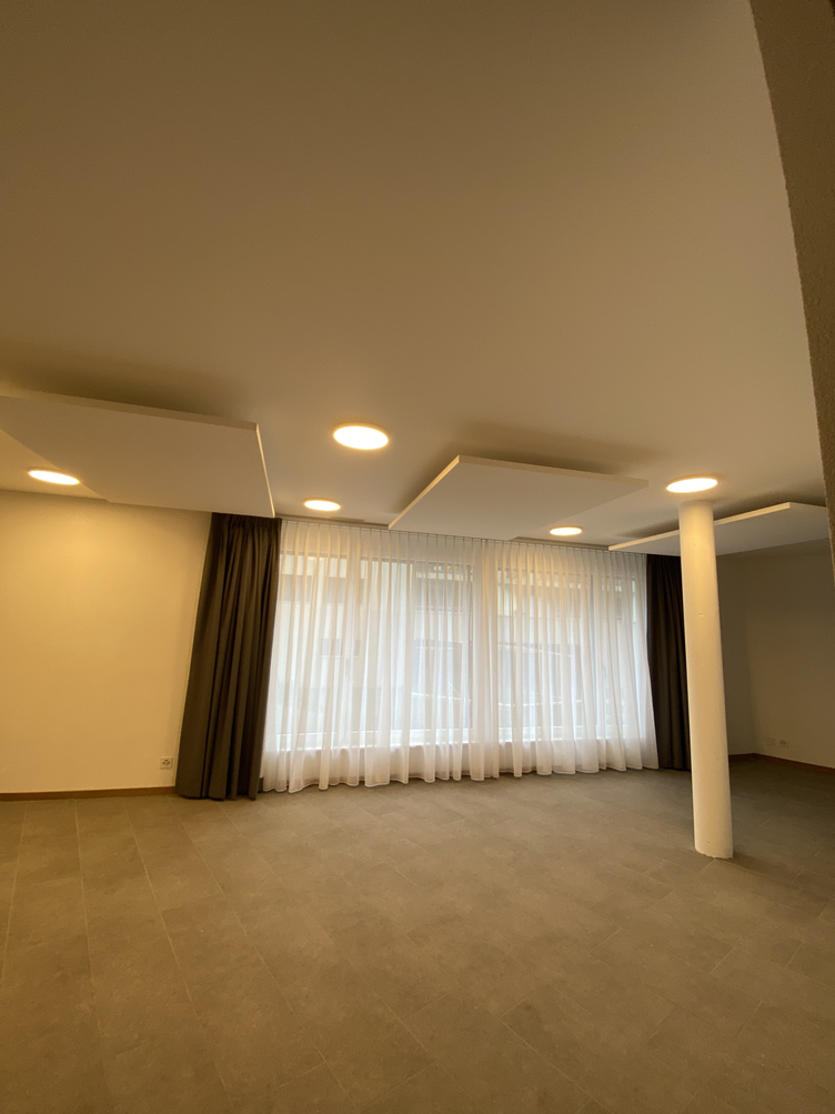 Pièce beige vide avec moquette, spots encastrés, une colonne blanche et de grandes fenêtres à rideaux