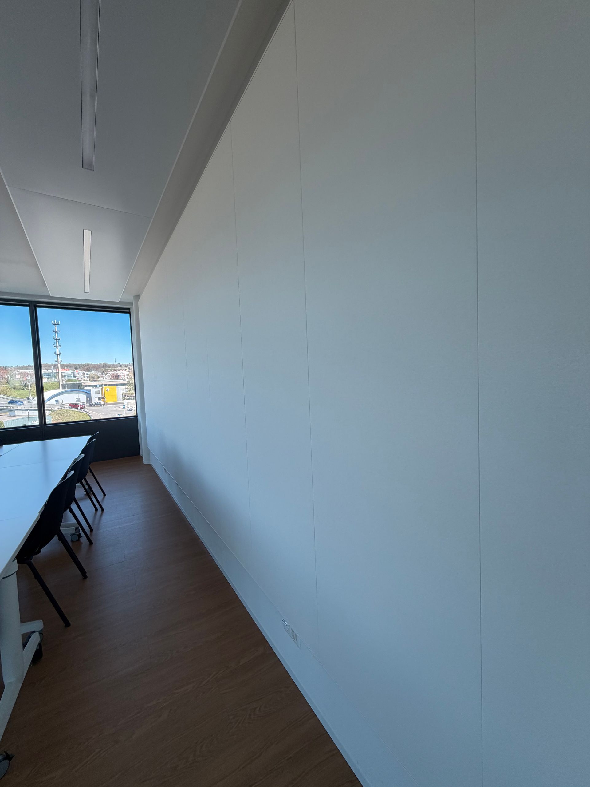 Espace de bureau long et étroit avec mur blanc, parquet, longue table et chaises près d'une fenêtre donnant sur la ville