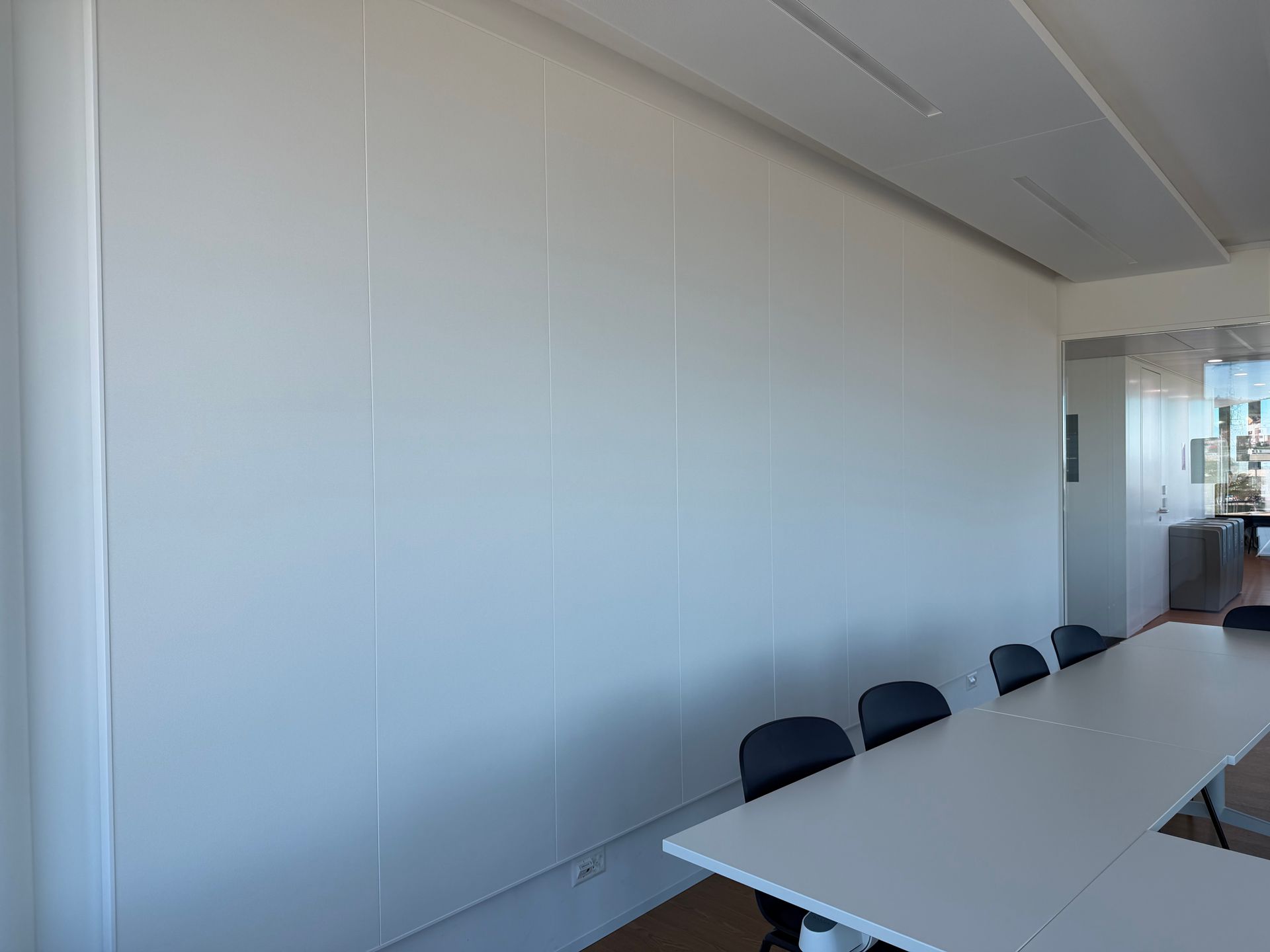 Salle de conférence aux murs blancs, avec une longue table et plusieurs chaises noires près d'une porte vitrée.