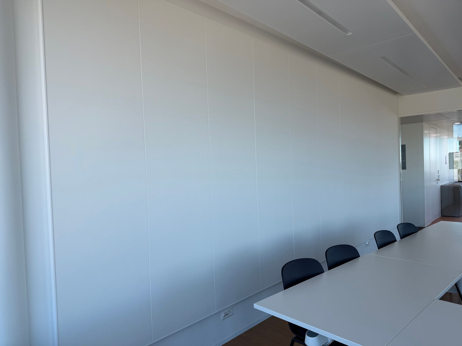 Salle de conférence vide avec murs blancs, longue table et chaises noires près d'une fenêtre.
