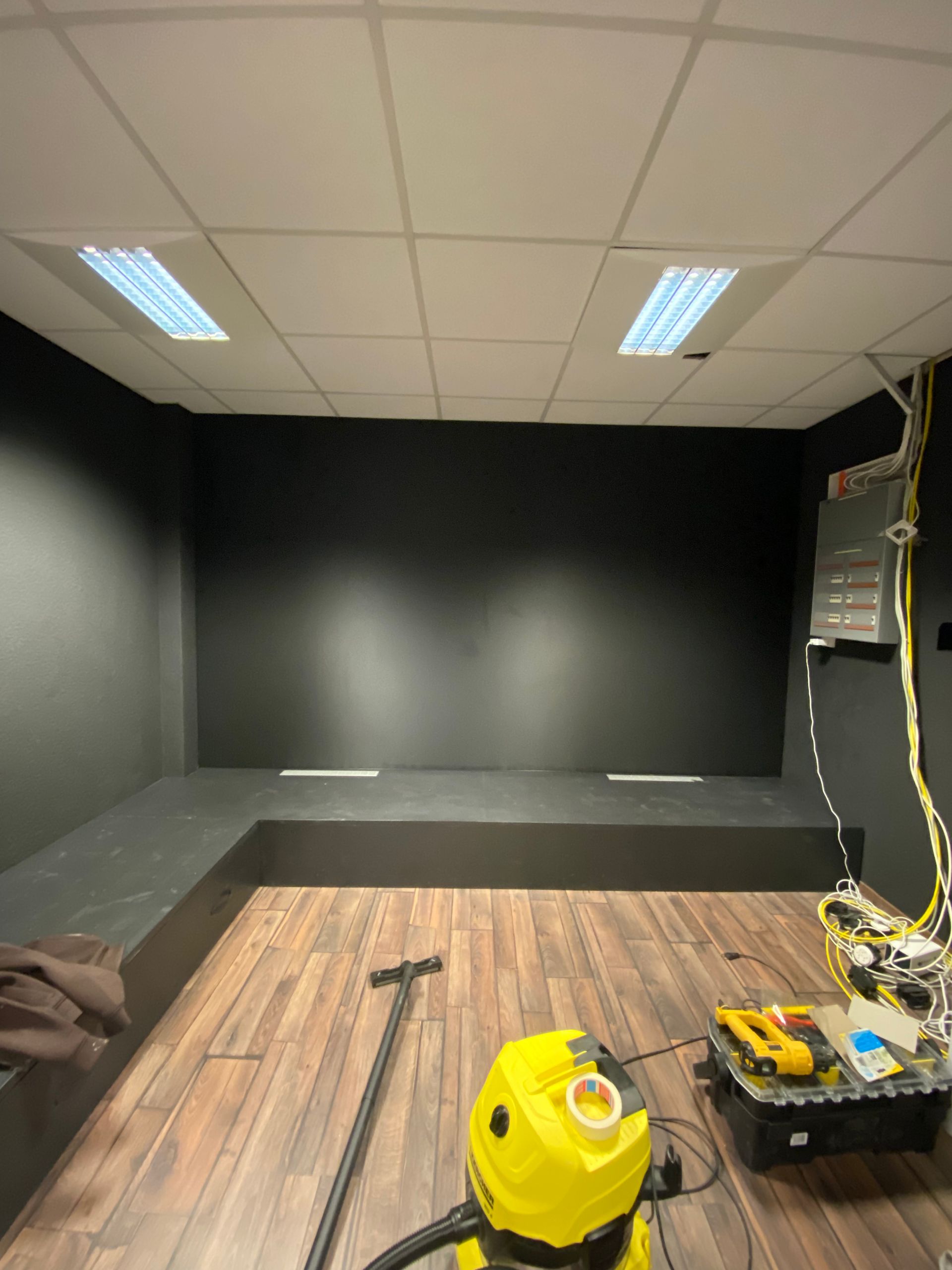 Pièce noire vide avec un sol en bois, des luminaires au plafond, des outils et un aspirateur d'atelier jaune au premier plan.