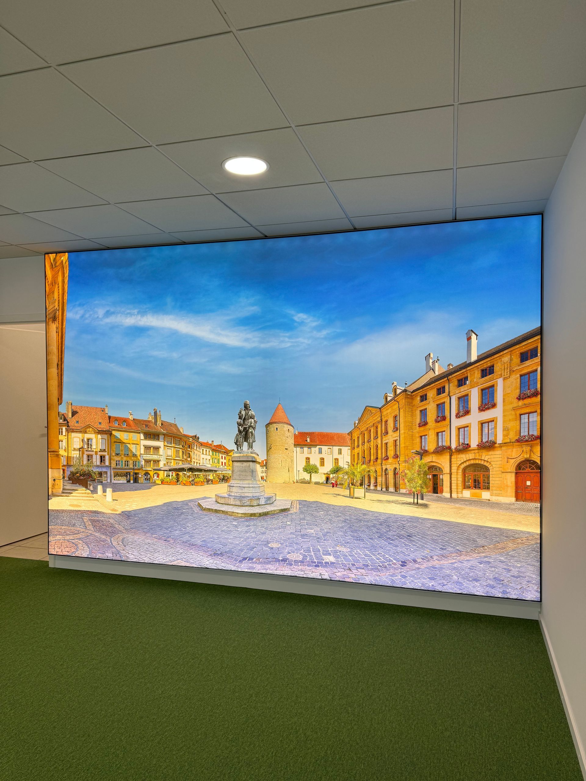 Écran intérieur diffusant une place de ville européenne ensoleillée avec des bâtiments colorés et un ciel bleu