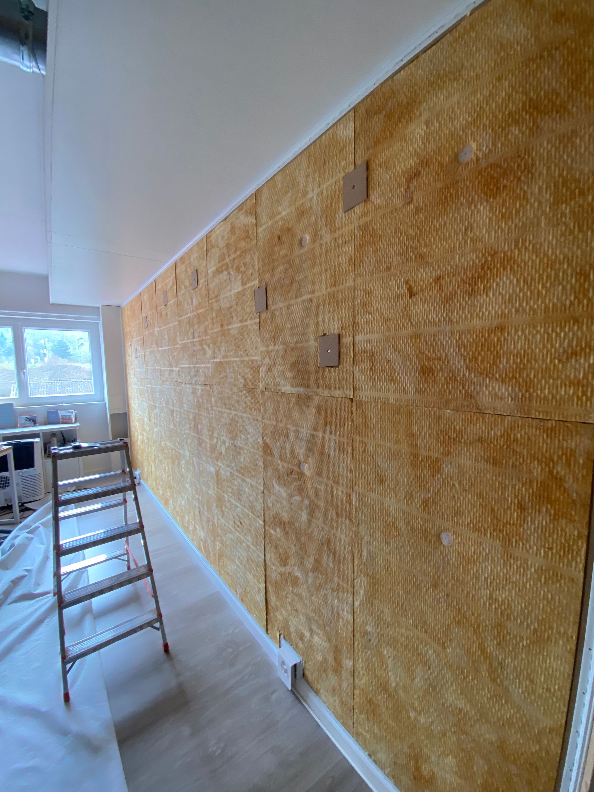 Un mur intérieur est recouvert d'un matériau texturé orange ; une échelle se dresse dans un couloir lumineux.