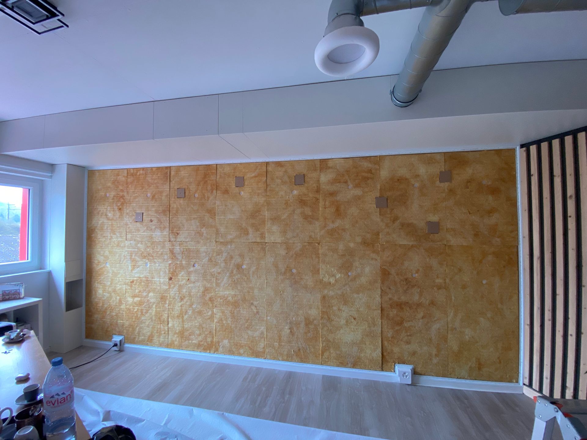 Pièce vide avec un grand mur d'accent beige, une fenêtre et des conduits d'aération apparents au plafond.