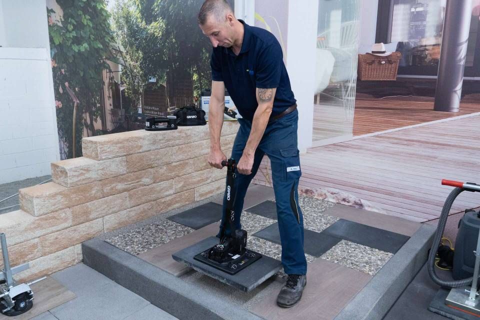 Ein Mann hebt mit einem Hebelwerkzeug eine schwere Steinplatte in einem Innenraum an. Zu sehen sind graue Steine ​​und Kies.