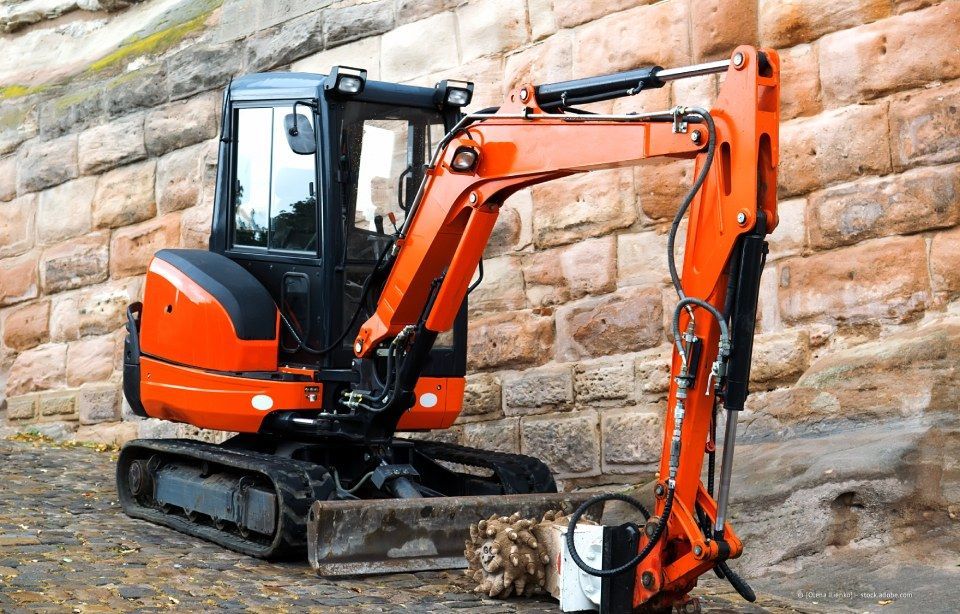 Ein orange-schwarzer Minibagger parkt neben einer Steinmauer.