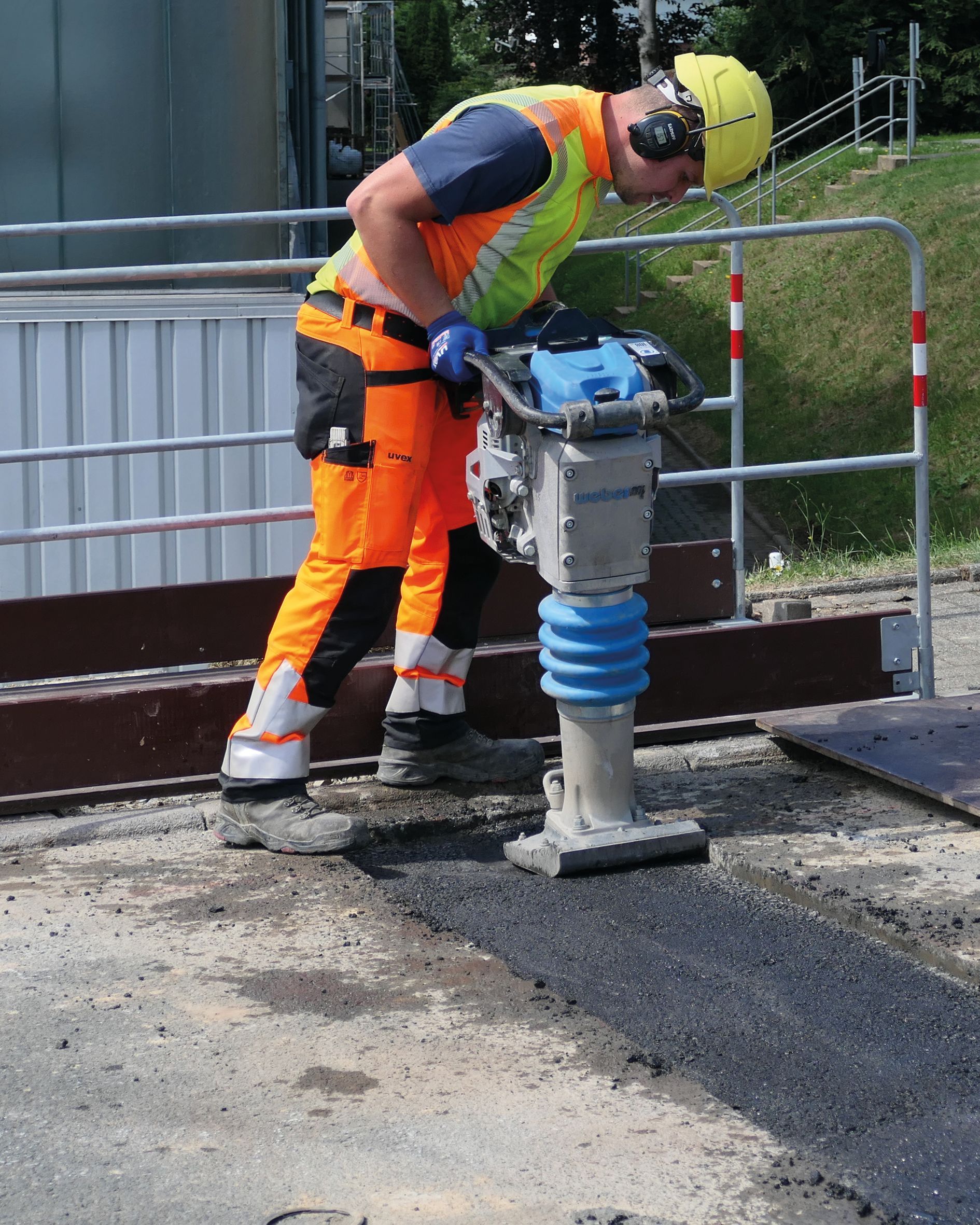 Ein Bauarbeiter bedient eine Walze auf Asphalt. Er trägt Schutzausrüstung, die Arbeit findet im Freien statt.