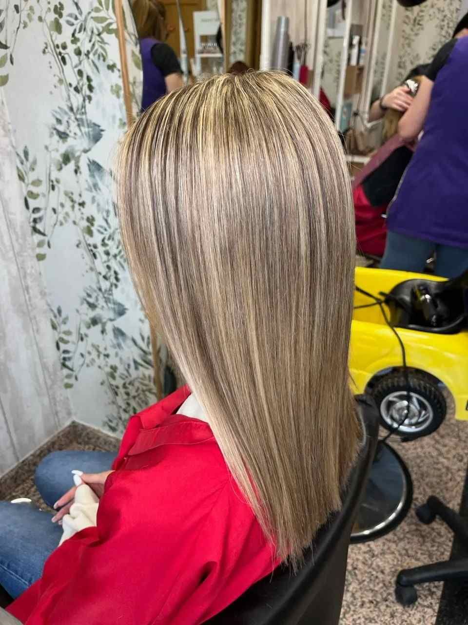 Mujer con largas trenzas con mechas, sentada en una silla de peluquería, con una capa roja, vista desde atrás.