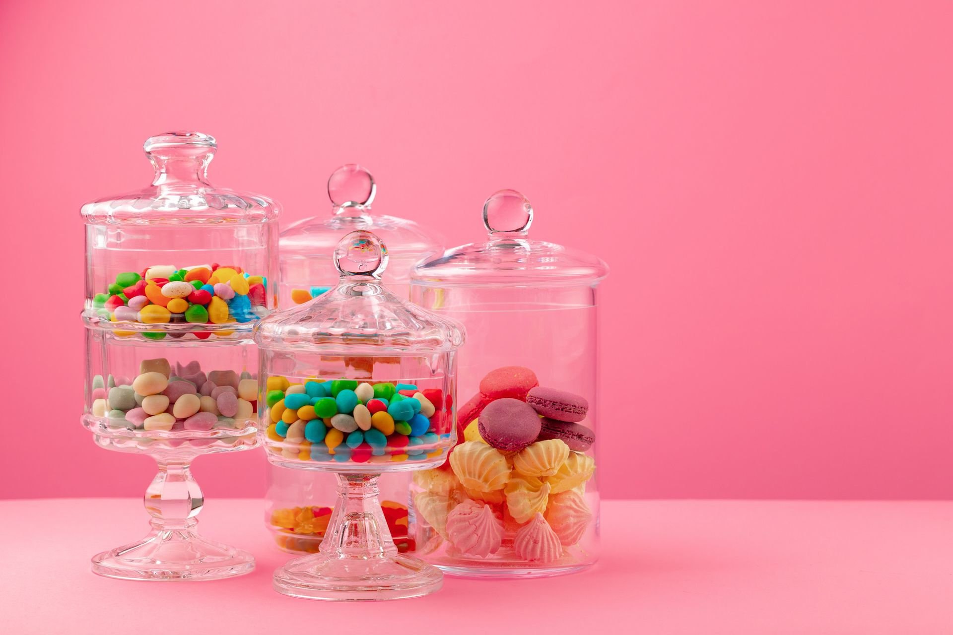 Tres frascos de vidrio llenos de diferentes tipos de dulces sobre un fondo rosa.