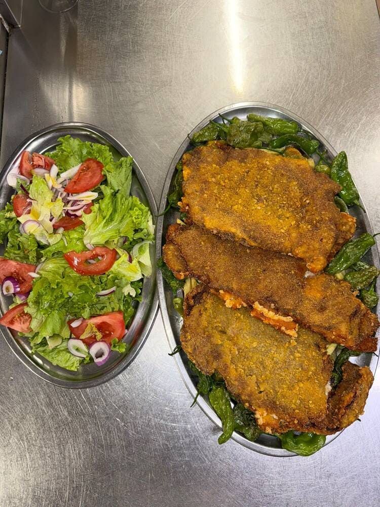 Dos bandejas metálicas con ensalada y chuletas empanadas fritas sobre un mostrador de acero inoxidable.