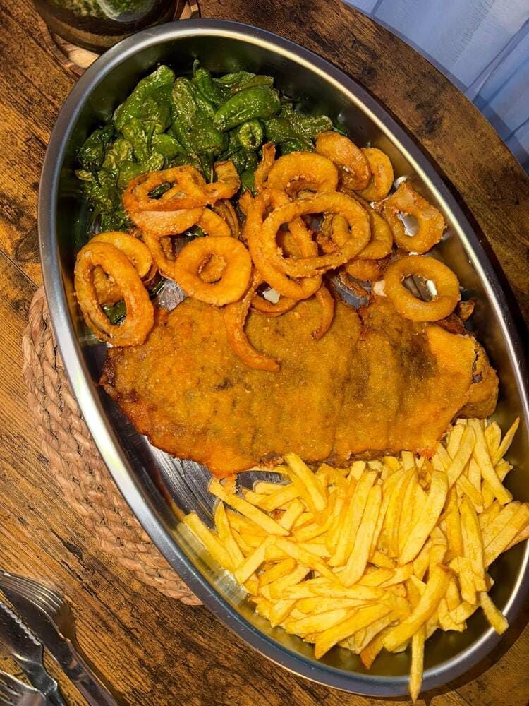 Vista aérea de una chuleta empanizada con papas fritas, aros de cebolla y verduras en un plato sobre una mesa de madera.