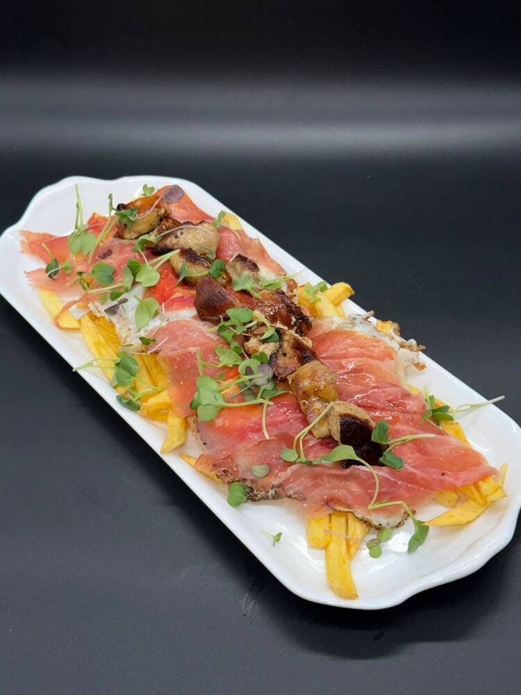 Plato rectangular de jamón serrano cortado en lonchas finas con verduras, queso y salsas sobre un fondo negro.
