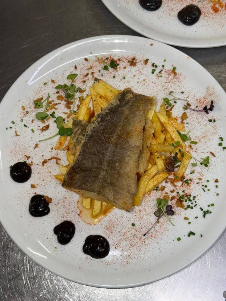 Filete de pescado emplatado sobre patatas fritas, adornado con hierbas y salsa oscura en un plato blanco en un ambiente de cocina.