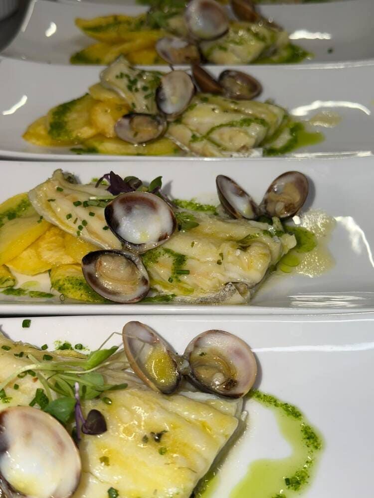 Filetes de pescado emplatados con almejas y salsa de hierbas verdes en platos blancos.