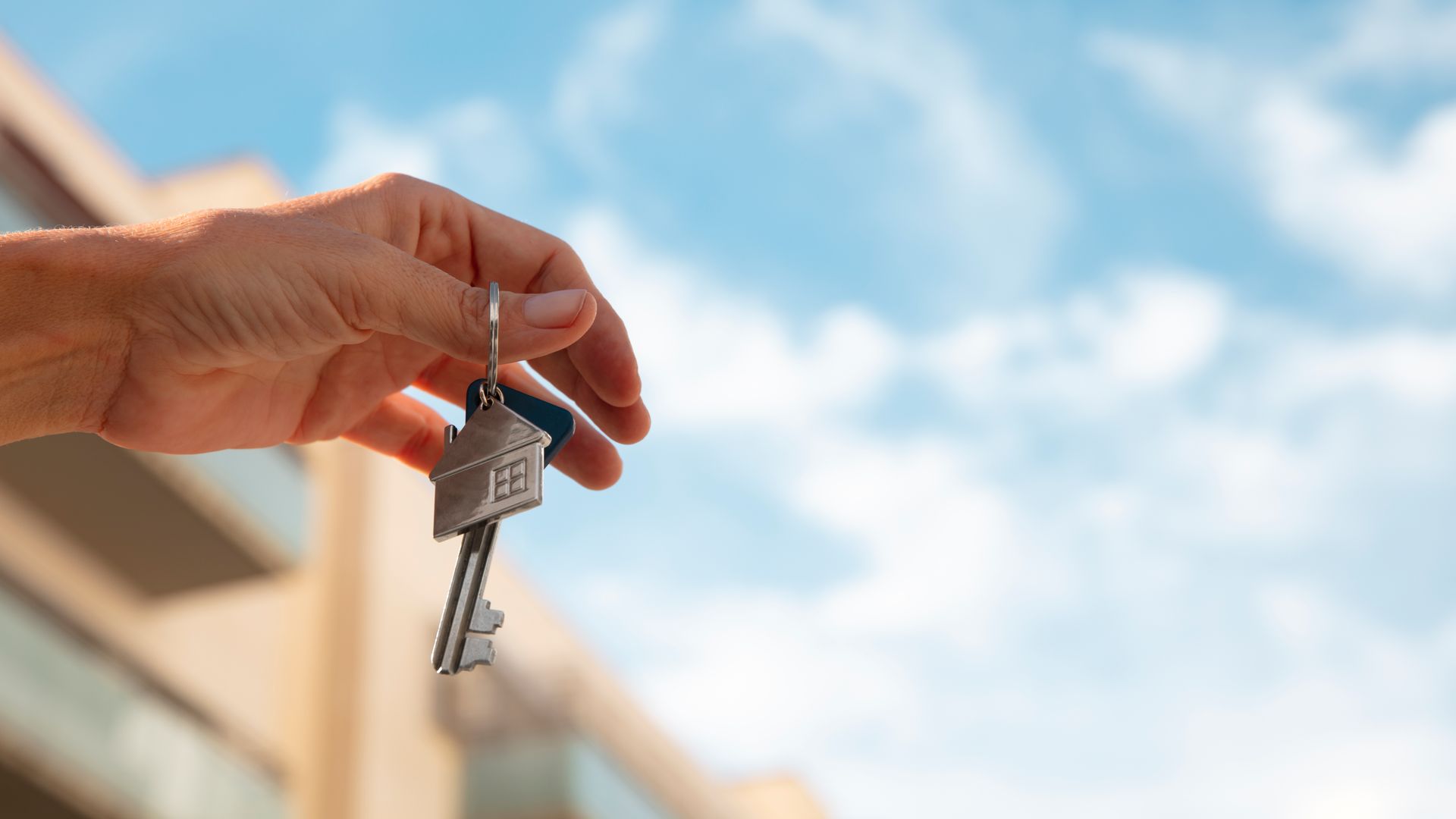 Mano sosteniendo las llaves con un llavero con forma de casa, contra un edificio borroso y un cielo.