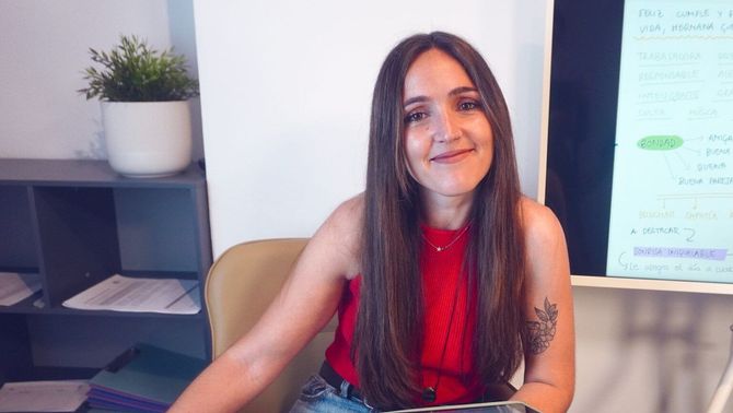 Mujer con una blusa roja sonriendo en un escritorio, con un portátil y una pizarra en una oficina luminosa
