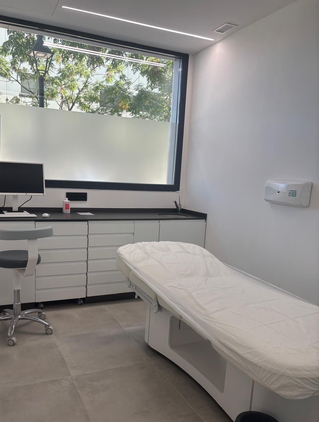 Sala de exploración médica con mesa de exploración, armarios, ventana y lavabo.