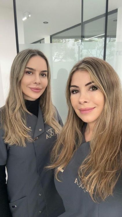 Dos mujeres con uniformes grises sonriendo, de pie en una habitación muy iluminada.
