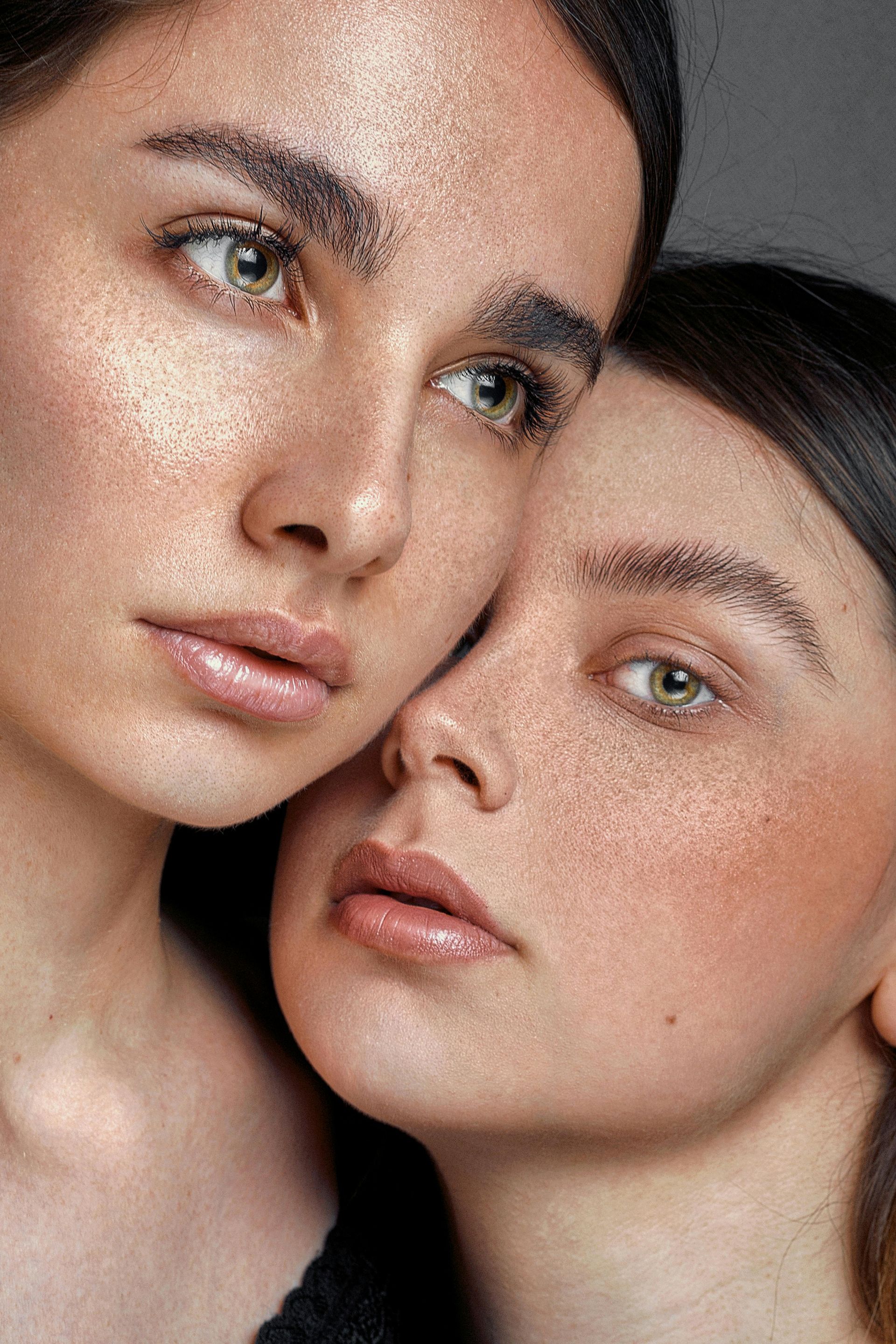 Dos mujeres con maquillaje brillante, mirando hacia la cámara, primer plano, fondo oscuro.